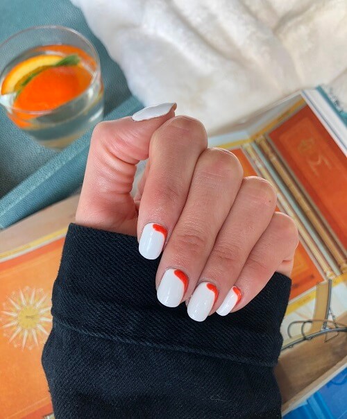 Single hand with black jacket cuff highlighing a white and orange nail art manicure by mobile professional Lauren Y